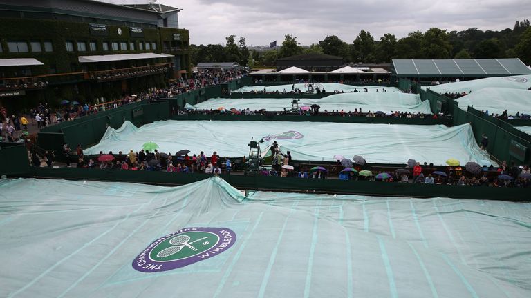 Spectators with umbrellas sit alongside outer courts with the rain covers on at Wimbledon