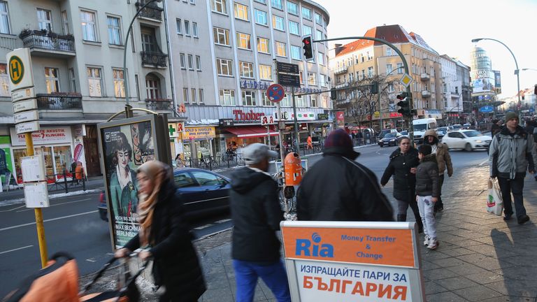 People walk along Karl-Marx-Strasse shopping street next to a sign advertising money transfers to Bulgaria in ethnically-diverse Neukolln, Berlin. 