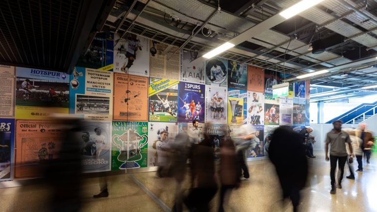 Supporters walk by a wall covered in large, poster-sized imagery of match day programmes at Tottenham Hotspurs' new stadium