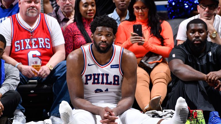 A shoeless Joel Embiid sits courtside during a Sixers game