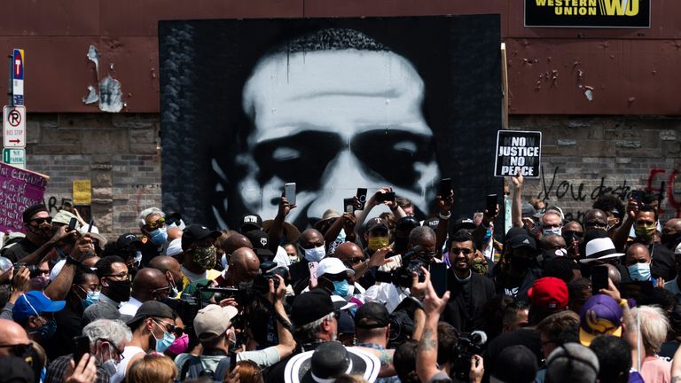 A painting of George Floyd stands behind a group of people gathered at a memorial on the block where he was killed by police