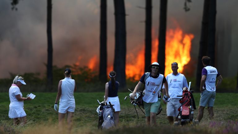 Players, Sophie Powell, Cara Gainer and Gabriella Cowley and their caddies look on after a fire behind the tenth hole stops play on day three of The Rose Ladies Series at Wentworth Golf Club