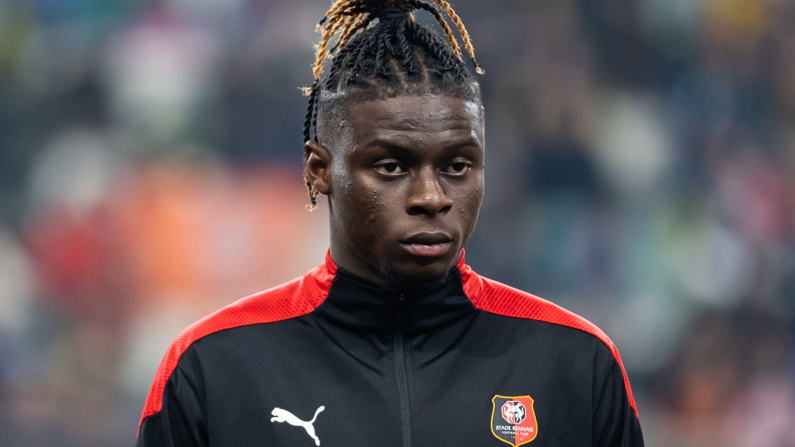  A young male football player with long braided hair and a black and red jersey with a white collar is shown from the chest up.