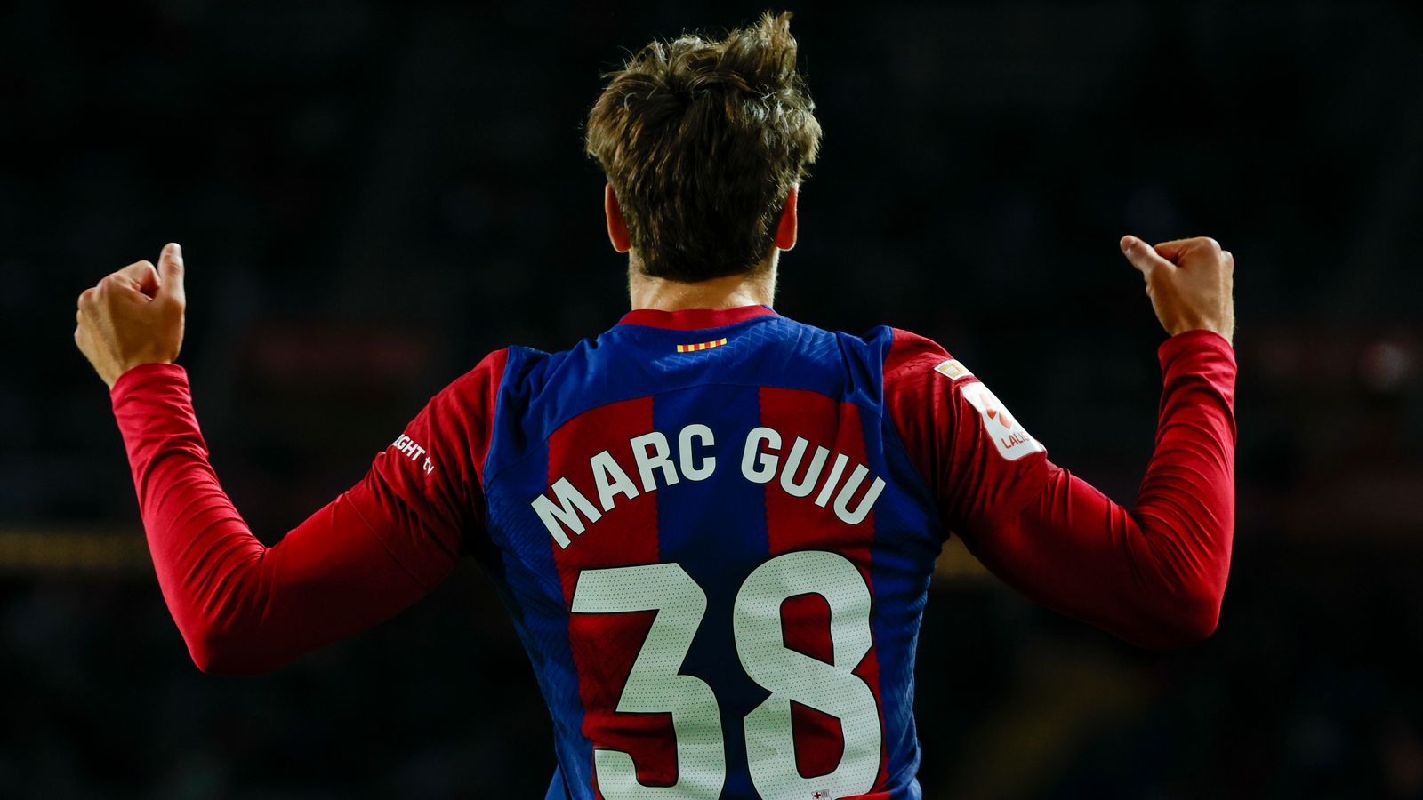  The image shows the back of a young male football player with his arms in the air celebrating with the text on his shirt saying 'Marc Guiu 38'. He is wearing a red and blue striped shirt with the name of the tournament 'European U17 Championship' on his sleeve.